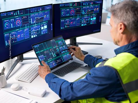 A man in coveralls sits at a desk with three screens monitoring a large graphic dashboard.